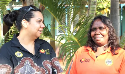 Two Indigenous women standing in front of a palm tree, one wearing a black shirt with Aboriginal design and the other an orange shirt.