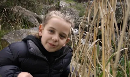 Young girl in a black jacket looks to camera. Beside her are yellow reeds and behind are rocks.