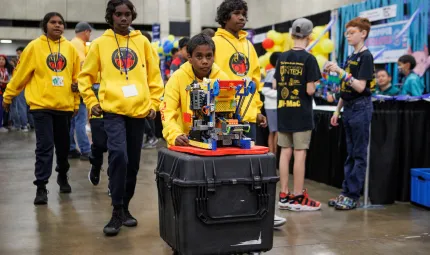 Four primary school aged Indigenous kids in yellow hoodies push a wheeled, black case with a robot on top. They're in a large hall surrounded by other kids.