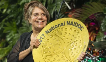 An Aboriginal woman holding a large golden coloured plate with the words “2017 National NAIDOC Lifetime Achievement Award”.