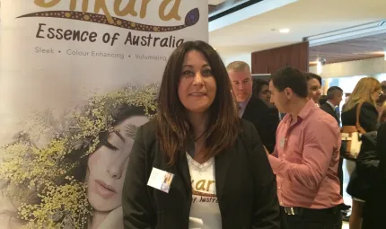 A woman, Julie Okley, stands in front of a poster displaying the words Dilkara, Essence of Australia. There are other people in the background.