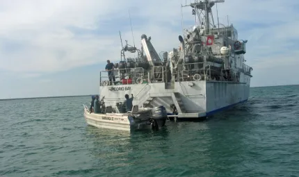 Small motor boat with Djelk rangers in water at back of naval vessel in open sea.