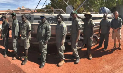 Group of men and a woman dressed in ranger uniforms stand in front of a boat. In the background are trees.