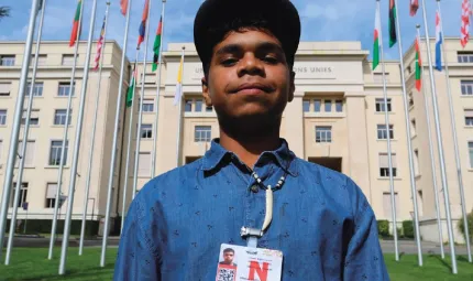 A young Aboriginal man in blue shirt and dark cap with a security pass around his neck stands in front of a large building. Immediately behind him are several flags on flagpoles.