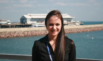 Woman dressed in black shirt stands in foreground with blue water and buildings in the background.