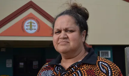 An Aboriginal woman with her hair up and wearing a polo shirt with Indigenous design stands in front of a building.