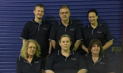 Two rows of people dressed in blue polo neck shirts in front of a blue roller door; three standing in the back row and three are sitting in the front.