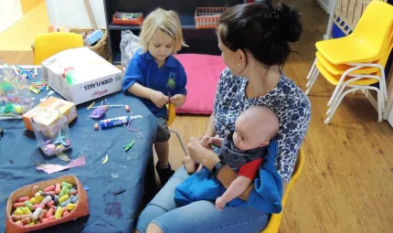 Woman in blue and white blouse sits at small table with craft materials. She holds a baby while looking at a young child in blue shirt working with craft material.