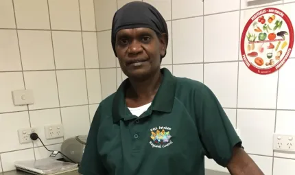 A middle-aged Indigenous woman in green shirt and head covering stands in front of a stainless steel bench with a white tiled wall in the background covered in powerpoints and a poster about fruit and vegetales.