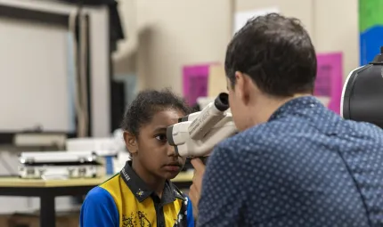 A young child looks into a piece of eye testing apparatus while a man looks into the other end. In the background is a table and other equipment.