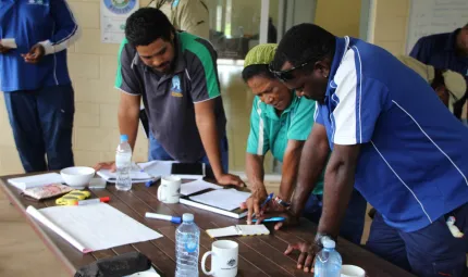 Three Torres Strait Islanders are standing at a desk looking at a note book. The woman in the middle is explaining something to the two men around her. There are other people in the background.