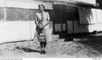 A woman wearing a uniform and hat, stands on grass in front of a building.