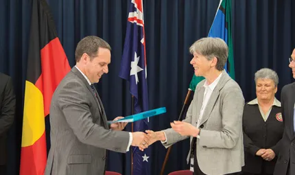 Group of people standing in front of Aboriginal, Australian and Torres Strait Islander flags, watching woman in bone grey coloured jacket shake hands with man in grey jacket.