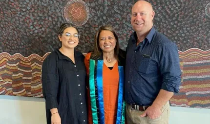 Three people stand shoulder to shoulder. At left is a woman in a dark dress. In the middle is another woman in an orange dress with a green, black and blue sash around her neck and at right is a man in a blue shirt and fawn trousers.