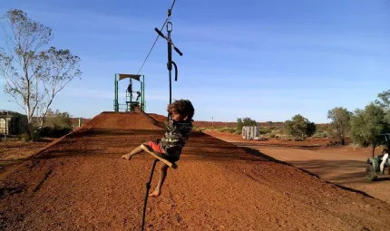 Aboriginal child on a cable suspended over brown soil. In the background is bare soil and some trees.