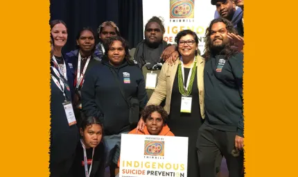 Group of youth and adults wearing mainly dark clothing stand or squat in a room. One holds a sign that says Indigenous Suicide Prevention. In the background is a large poster, some dark curtains and some grey/green curtains.