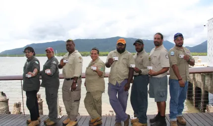 Line of Indigenous men and women dressed in ranger uniforms display their individual ranger accreditation. In the background is a wharf, a bay and a hill.