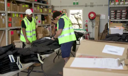 Two men wearing high-visibility vests and blue shorts working in a warehouse filled with boxes. In front of the two men are tables piled with jackets and work clothing.