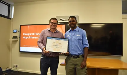 Two men stand side by side and hold between them a large framed certificate. The man on the left wears a checked shirt and dark trousers. The man at right wears a blue shirt and green trousers. They stand in a room with two large TV screens in the back.