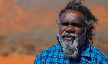 Head and shoulders shot of Aboriginal man with greying beard and wearing a blue checked shirt. In the background is an arid and ochre coloured landscape.