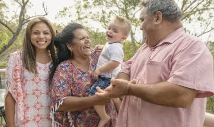 Three generations of an Indigenous family. At left is a young adult woman in pale dress. Next to her is an older woman in blue and orange dress holding a young boy in shorts and white t-shirt. At right is a man in pale pink shirt. Trees in the background.