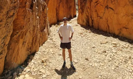 A man in light coloured shirt and dark shorts, wearing a cap and sunglasses, stands on rocky ground in a gorge with ochre coloured walls.