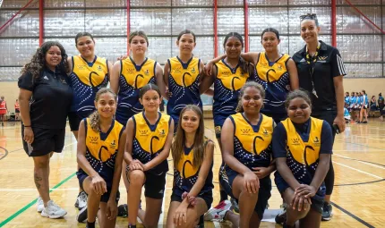 Two adult women in blue flank 10 young women in yellow and blue sports uniforms as they stand or kneel on a basketball court. In the background is a shiny stadium wall.