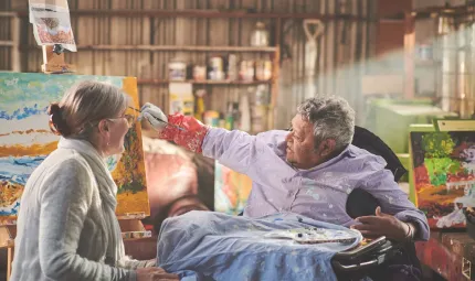 A woman and a man sit and talk to each other as the man paints a landscape picture. In the background is a wall with paints and other apparatus on shelving.