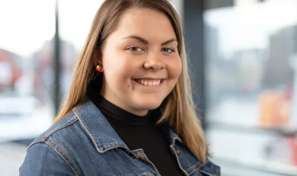 Head and shoulders shot of a young adult woman with long brown hair and wearing a dark top and denim jacket.