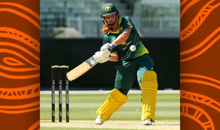 Young woman dressed in green cricket helmet and clothing and with yellow pads on her legs, swings a cricket bat at a white ball on a grass covered playing arena. In the background is a dark wall and seating above that.