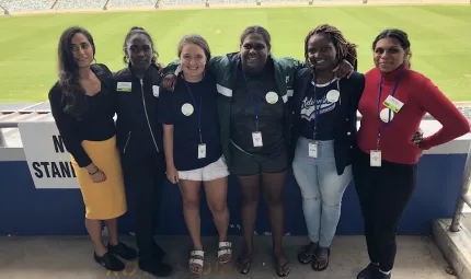 Six Indigenous and non-Indigenous young women in different coloured and styles of clothing stand arm in arm. In the background is a playing field.