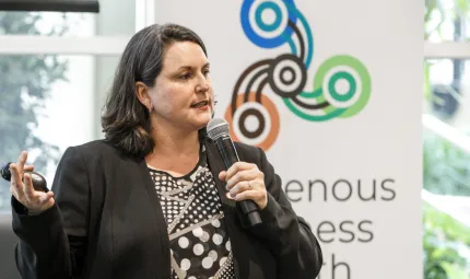 A woman with brown hair and wearing a brown jacket speaks into a microphone. In the background is a large poster in front of a window behind which are trees. The poster has a logo featuring three sets of concentric circles linked by curved lines.