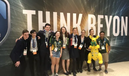 Line of young people arm in arm wearing various clothing styles stand on a hard pale surface and in front of a wall with the words in very large print: Think Beyond.