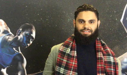 A young aboriginal man wearing a tartan scarf stands in front of a picture of a female dancer.