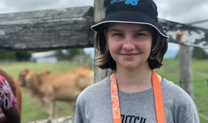 A young girl wearing a grey t-shirt and black hat, looking at the camera. She is standing in front of a paddock with cows.