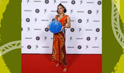 A smiling woman stands on a red carpet, holding a large trophy which is round and vibrant blue with gold writing. It is shaped like a plate. She is standing in front of a wall covered in NAIDOC logos.