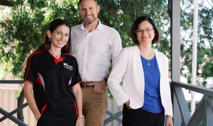 Three people stand on a porch facing the camera. At left is a woman in dark shirt. Next to her is a man in light shirt and brown trousers and next to him is a woman in white jacket, blue top and dark pants. In the background are trees and a fence.