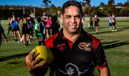 A man standing on a football field wearing a black and red polo shirt holding a yellow football. There are players behind him wearing different coloured uniforms.