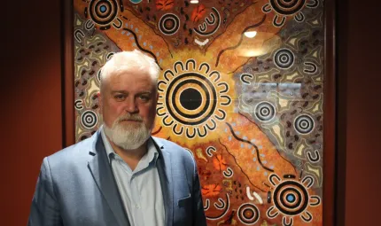 Aboriginal man with white beard and hair, dressed in blue suit and shirt stands in front of artwork of Indigenous design featuring circles, horseshoe shapes and squiggly lines.