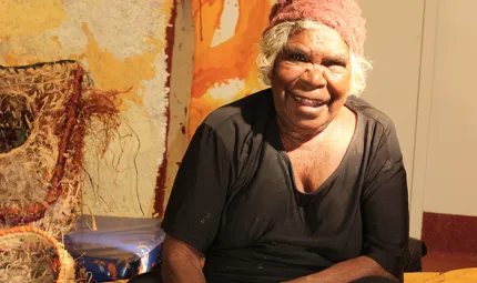 An elderly woman in black dress and reddish beanie is sitting down. In front of her is artwork featuring some birds. In the background is more art and a beige coloured wall.