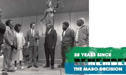 Five Indigenous men, one Indigenous woman and one non-Indigenous man stand in half circle in front of a statue of a woman holding a sword in the right hand and a set of scales in the left.  Words: 25 years since The Mabo Decision Remember