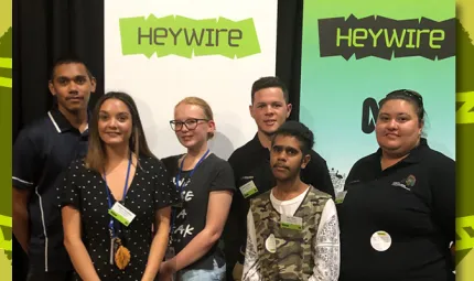 Six young people stand in front of two pull-up banners.The banners both have the word ‘Heywire’ written on them.