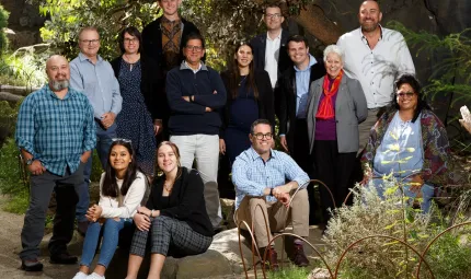 Group of people in various dress types face the camera. Some stand and others sit on rocks. In the background is a tree and other foliage. In the foreground is a fence, rock and plants.