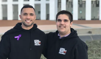 Two young Indigenous men dressed in black hoodies stand arm in arm in front of the white pillars of the Australian Parliament House. The text on their jackets read National Indigenous Youth Parliament.