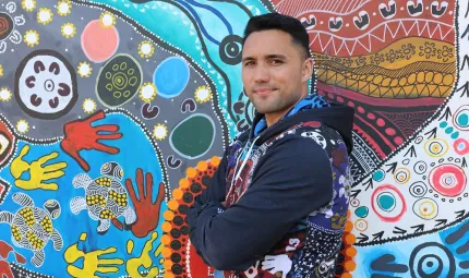 A young adult man in a mainly blue top stands in front of a painting of hands, circles, animals and other features. The painting has blue, grey and red background colours.