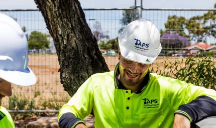 Two men in white hard hats and green work wear tops stand near each other. In the background is a tree, a fence and buildings in the distance.