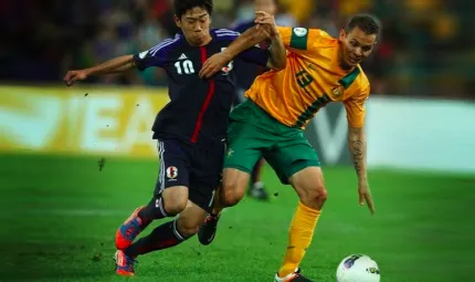 Two men struggle for possession of a soccer ball. The man on the left is in blue and the man on the right is in a yellow top and green shorts.