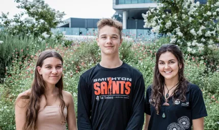 There are 3 young Indigenous people facing the camera and smiling. They are standing shoulder to shoulder, outside on an overcast day, in front of flowering grasses and trees.