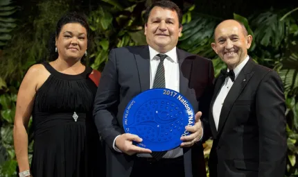 Three people standing together with plants behind them. The man in the middle is holding a large blue plate while a woman is standing to his right and man to his left.