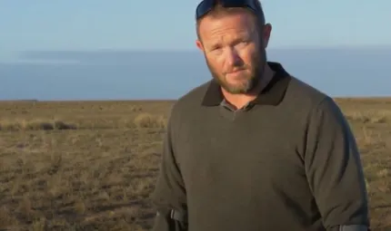 A middle-aged man in dark jumper and sunglasses on his head stands, supported by crutches, on an open plain. In the background and above is blue sky.